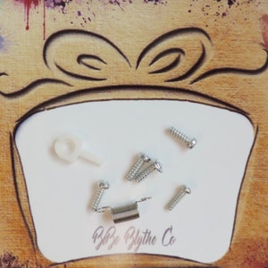 May include: A close-up of a white surface with a silver spring, a white plastic hook, and six silver screws. The surface is surrounded by a brown wooden frame with a painted design of a gift box with a bow.