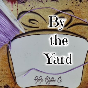 Puede incluir: Una bobina de cordón de color lavanda con el texto "By the Yard" y "Byp Blythe Co" sobre un fondo blanco.