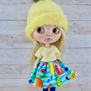 May include: A doll wearing a yellow knitted hat with a green leaf on top, a yellow and white striped shirt, a blue and white skirt with fruit designs, blue tights, and black shoes.
