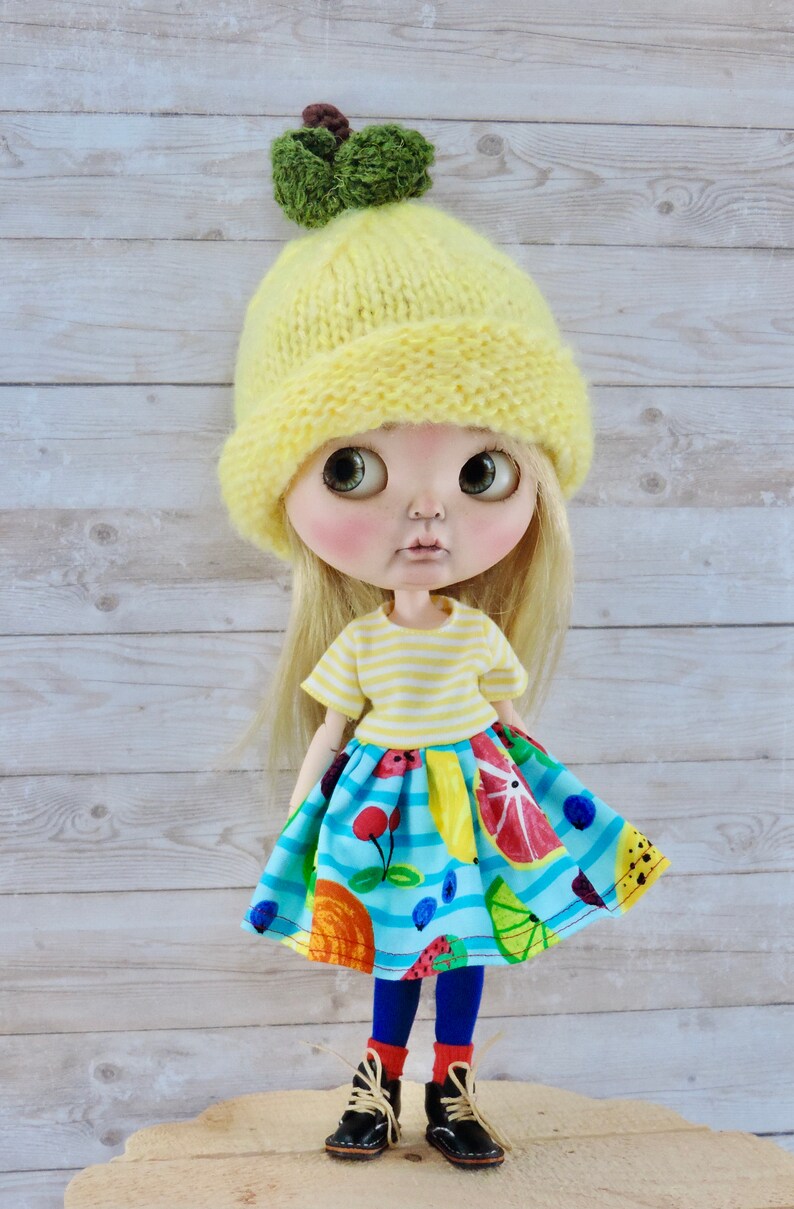 May include: A doll wearing a yellow knitted hat with a green leaf on top, a yellow and white striped shirt, a blue and white skirt with fruit designs, blue tights, and black shoes.