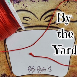 May include: A spool of bright red cord with the text "By the Yard" and a butterfly illustration. The cord is unwound, suggesting it is sold by the yard. The background has a vintage, artistic design.