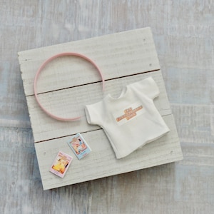 May include: A white t-shirt with the text "The Extraordinary One" on the front, a pink headband, and two small books with colorful covers on a white wooden surface.