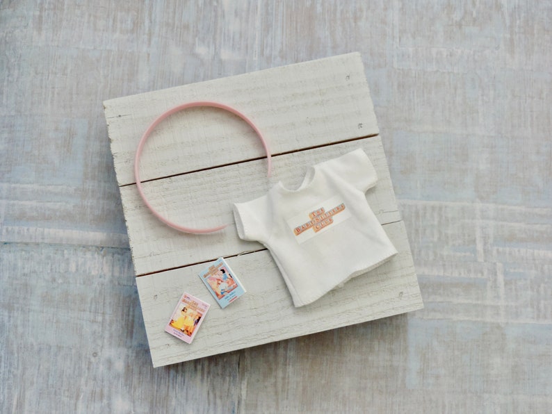May include: A white t-shirt with the text "The Extraordinary One" on the front, a pink headband, and two small books with colorful covers on a white wooden surface.