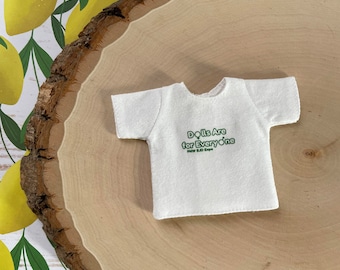 May include: White t-shirt with the text "Dolls Are for Everyone" in green lettering on a wood background with a lemon pattern.