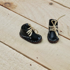 May include: Two miniature black leather boots with white stitching and beige laces on a light wood background.