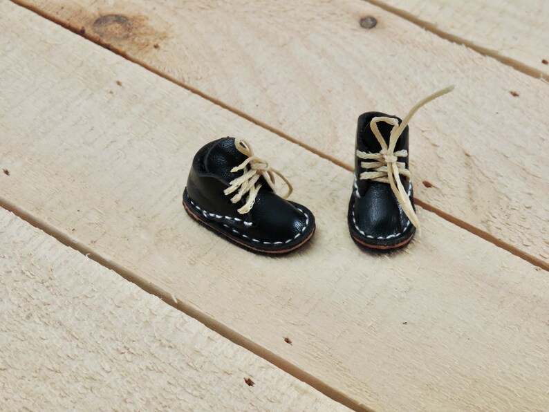 May include: Two miniature black leather boots with white stitching and beige laces on a light wood background.