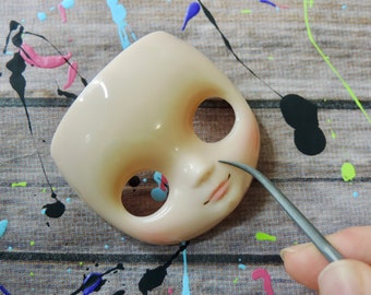 May include: A close-up of a doll's head with a pair of tweezers being used to apply a detail to the doll's nose. The doll's head is made of a light-colored plastic and has a painted-on face with closed eyes and a pink mouth.