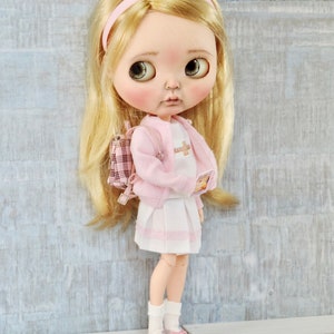 May include: A doll wearing a pink cardigan, white skirt, and pink shoes. The doll has blonde hair and is holding a book. The doll is wearing a pink headband and has a pink backpack.