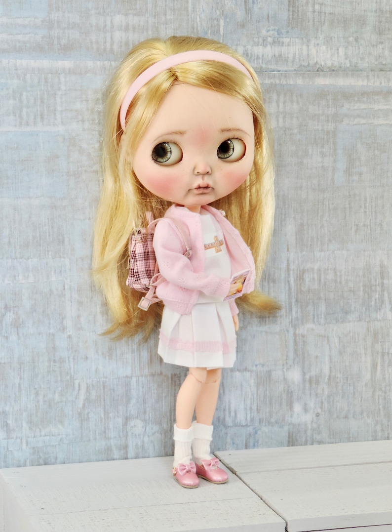May include: A doll wearing a pink cardigan, white skirt, and pink shoes. The doll has blonde hair and is holding a book. The doll is wearing a pink headband and has a pink backpack.