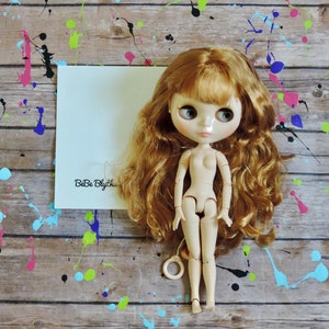 May include: A nude-colored fashion doll with long, wavy auburn hair and large, expressive eyes. The doll is positioned in front of a white card with the text "BeBe Blythe" on a wooden surface with colorful paint splatters.