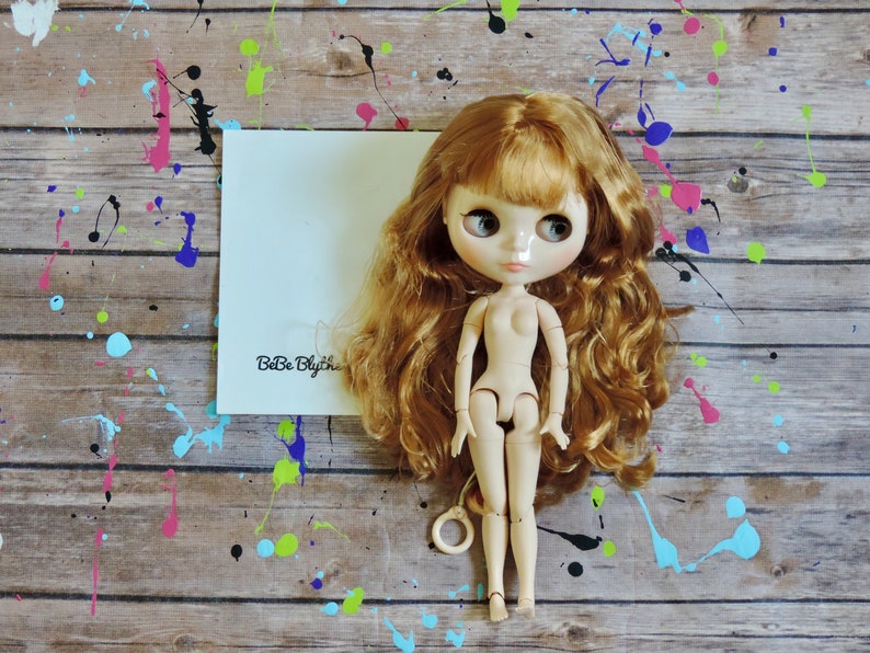 May include: A nude-colored fashion doll with long, wavy auburn hair and large, expressive eyes. The doll is positioned in front of a white card with the text "BeBe Blythe" on a wooden surface with colorful paint splatters.