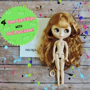 May include: A nude-colored fashion doll with long, wavy auburn hair. The doll has large, expressive eyes and is articulated. A green starburst-shaped sign reads "4 Free Eye Chips with Doll Purchase!" The background is a wood-grain surface with paint splatters.