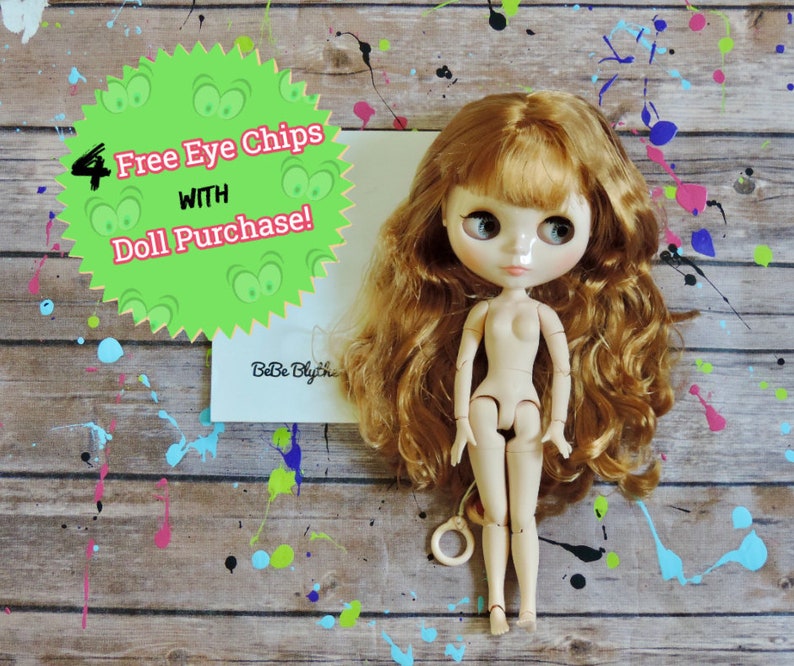 May include: A nude-colored fashion doll with long, wavy auburn hair. The doll has large, expressive eyes and is articulated. A green starburst-shaped sign reads "4 Free Eye Chips with Doll Purchase!" The background is a wood-grain surface with paint splatters.