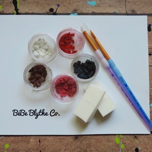 May include: A collection of art supplies on a white surface, including small clear containers filled with colorful materials, two paintbrushes, and two small white sponges. The text "BeBe Blythe Co." is printed on the surface.