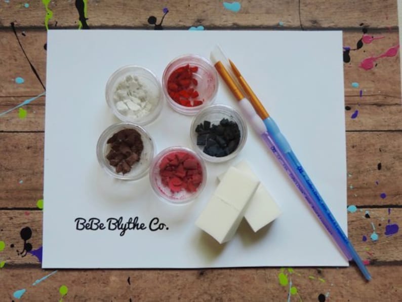 May include: A collection of art supplies on a white surface, including small clear containers filled with colorful materials, two paintbrushes, and two small white sponges. The text "BeBe Blythe Co." is printed on the surface.