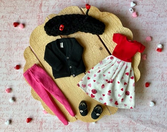 Könnte beinhalten: Ein Puppen-Outfit-Set mit einem schwarzen Cardigan, rosa Leggings, einem roten T-Shirt mit kurzen Ärmeln, einem weißen Rock mit rosa Herzen und schwarzen Schuhen.