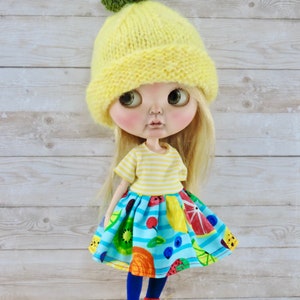 May include: A doll wearing a yellow knitted hat with a green pom-pom, a yellow and white striped shirt, a blue skirt with a fruit print, blue tights, and black shoes.