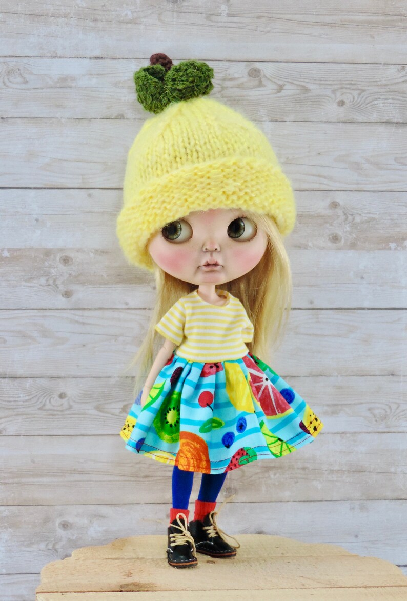 May include: A doll wearing a yellow knitted hat with a green pom-pom, a yellow and white striped shirt, a blue skirt with a fruit print, blue tights, and black shoes.