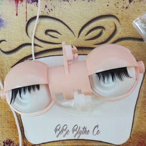 May include: A pair of pink doll eyes with black eyelashes, attached to a white string. The eyes are in a plastic holder with a white label that says "Baby Blythe Co."