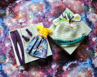 Könnte beinhalten: Ein Set Puppenkleidung mit einem gelben und blauen Kleid mit Einhorn-Druck, einer lila Leggings, zwei Paar lila Stiefeln und einer weißen Häkelmütze mit einem Einhorn-Horn.
