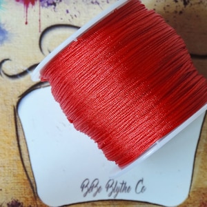 Puede incluir: Un carrete de cordón rojo brillante, enrollado firmemente en una bobina de plástico blanco. La bobina está en ángulo, revelando la textura del cordón. El fondo es un diseño artístico colorido con salpicaduras de pintura y el texto "Blythe Co".