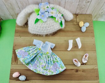 Könnte beinhalten: Ein Puppenoutfit mit einem blau-weiß gemusterten Kleid, einem blau-weiß gestreiften Oberteil, weißen Socken und weißen Schuhen. Das Outfit liegt auf einer Holzoberfläche mit einem weißen Hasenkopf und Ostereiern.