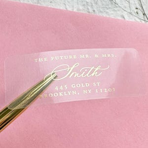 May include: A clear rectangular address label with gold foil text reading THE FUTURE MR. & MRS. Smith 445 GOLD ST BROOKLYN, NY 11201, being lifted by a gold-toned metal tool from a pink envelope surface.