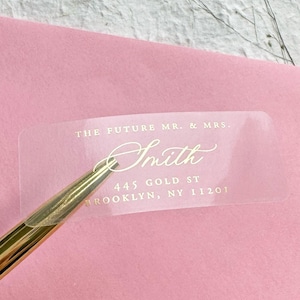May include: A clear sticker with gold lettering that reads "THE FUTURE MR. & MRS. Smith 445 GOLD ST BROOKLYN, NY 11201" is placed on a pink envelope. A gold pen is in the foreground.