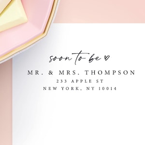 May include: A wedding invitation design with a pink and white background. The text "soon to be" is written in a script font, followed by the names "MR. & MRS. THOMPSON" and an address. A pink plate with gold trim and white and yellow treats is in the upper left corner.