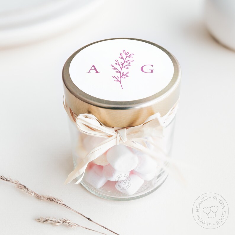 May include: A small clear glass jar filled with white marshmallows, featuring a gold-colored metal lid. The lid has a white circular label printed with the letters A and G flanking a pink botanical sprig. A light-colored raffia ribbon is tied around the neck.