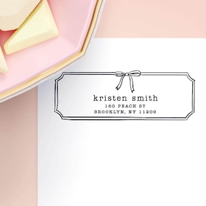May include: A white card with black text that reads "kristen smith" and the address "160 peach st brooklyn, ny 11209". The card has a decorative bow at the top and is in a black outlined frame.