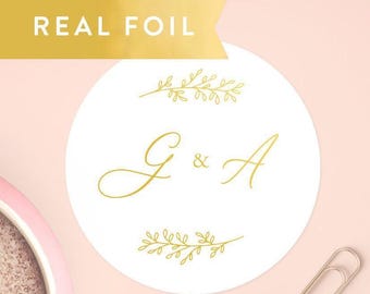 May include: A circular white sticker featuring gold foil lettering with the initials G & A in a cursive script, flanked by two horizontal gold foil botanical leaf sprigs. The text REAL FOIL appears in a gold banner at the top of the image.