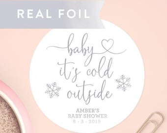 Könnte beinhalten: Ein runder weißer Aufkleber mit silbernem Folientext, der "baby it's cold outside" in Schreibschrift zeigt, ergänzt durch ein Herz und zwei Schneeflocken-Symbole. Darunter steht "AMBER'S BABY SHOWER 8-3-2019" in einer einfachen Schriftart.