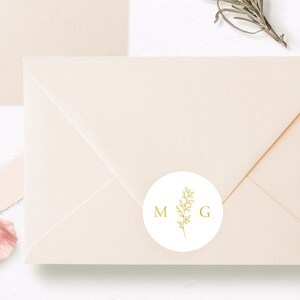 May include: A light peach-colored envelope sealed with a circular white sticker. The sticker features a gold-colored line drawing of a leafy sprig positioned between the capital letters M and G, printed in a simple, clean serif font.