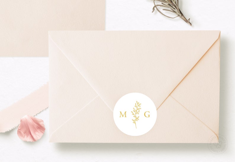 May include: A light peach-colored envelope sealed with a circular white sticker. The sticker features a gold-colored line drawing of a leafy sprig positioned between the capital letters M and G, printed in a simple, clean serif font.