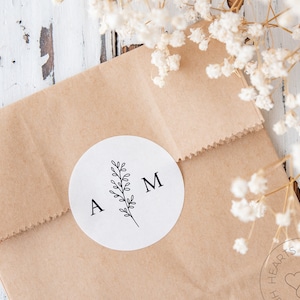 May include: A circular white sticker featuring the black printed letters A and M separated by a central line drawing of a leafy botanical sprig. The sticker is adhered to a light brown paper bag with a serrated top edge and a folded closure.