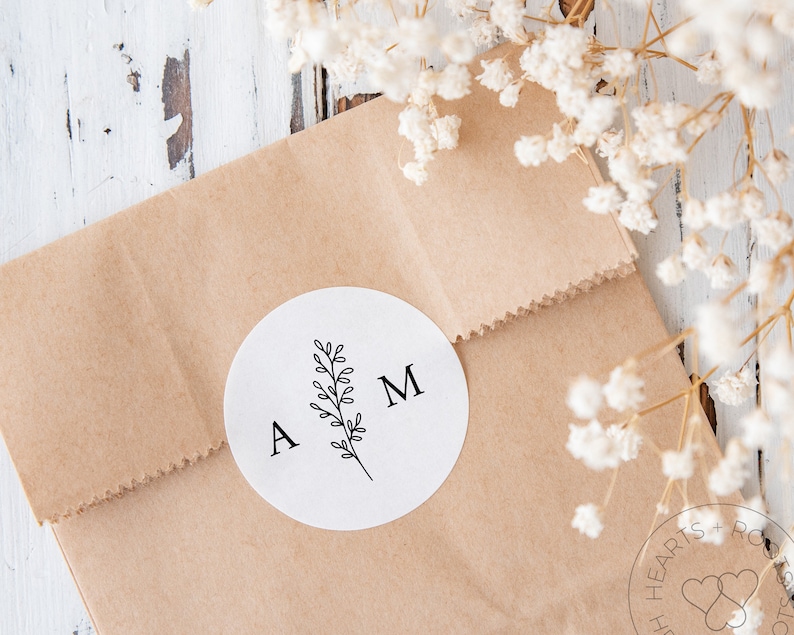 May include: A circular white sticker featuring the black printed letters A and M separated by a central line drawing of a leafy botanical sprig. The sticker is adhered to a light brown paper bag with a serrated top edge and a folded closure.