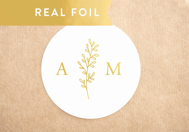 May include: A circular white sticker featuring a gold foil design with the letter A on the left, the letter M on the right, and a central botanical sprig graphic. The text REAL FOIL appears in white on a gold banner at the top of the image.