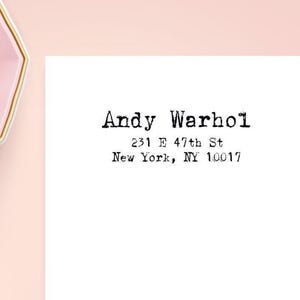 May include: A white rectangular card featuring black typewriter-style text that reads Andy Warhol, 231 E 47th St, New York, NY 10017. Beside the card is a light pink hexagonal bowl filled with pale yellow and light pink geometric shapes.