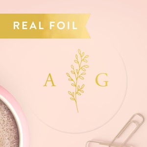May include: A circular sticker with a light pink background featuring the gold foil letters A and G separated by a gold foil botanical sprig. A gold banner at the top displays the text REAL FOIL. A metal paperclip is visible in the corner.