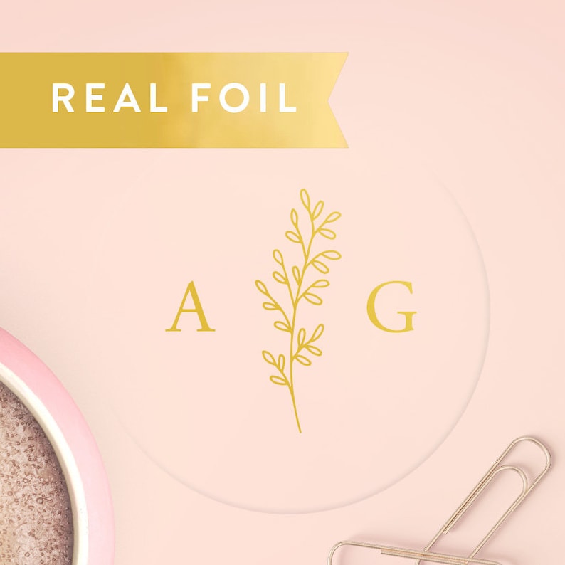 May include: A circular sticker with a light pink background featuring the gold foil letters A and G separated by a gold foil botanical sprig. A gold banner at the top displays the text REAL FOIL. A metal paperclip is visible in the corner.