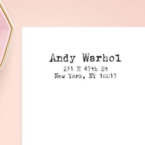 May include: A white rectangular card featuring black typewriter-style text that reads Andy Warhol, 231 E 47th St, New York, NY 10017. To the left, a light pink octagonal bowl contains several pale yellow and light pink geometric shapes.