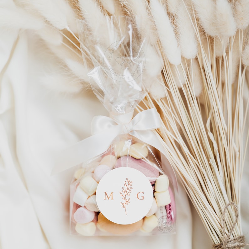 May include: A clear plastic favor bag filled with assorted pastel marshmallows and macarons, tied with a white satin ribbon. A circular white sticker on the front features the letters M and G in copper, separated by a delicate botanical illustration.