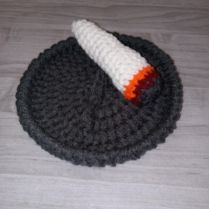 Puede incluir: Un artículo hecho a mano con ganchillo. Presenta una base circular gris oscuro con un elemento en forma de cuerno blanco, naranja y rojo. El artículo es probablemente una pieza decorativa o una creación artesanal única.