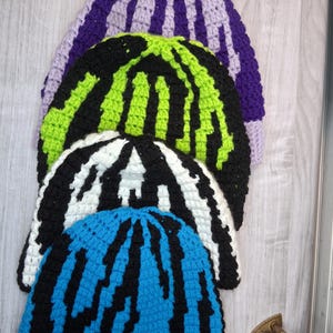 May include: Four handmade crochet beanies in a stack. The beanies are in various colors, including purple, black and green, black and white, and blue with black stripes. The top beanie has a purple pom-pom.