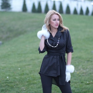 May include: A woman wearing a black button-down shirt with a pearl necklace and white fur gloves. She is standing in a grassy field.