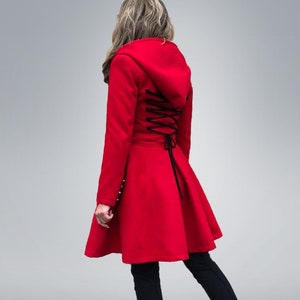 May include: A bright red hooded coat with a flared skirt and black corset-style lacing down the back. The coat features long sleeves and is paired with black leggings and sneakers.