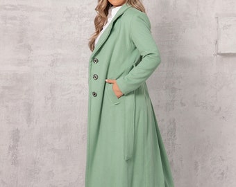 May include: A long, light green coat with a belt and button closure. The coat is worn by a person with long blonde hair and is standing on a gray concrete background.