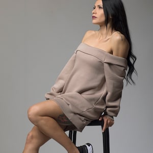 Puede incluir: Un vestido sudadera color topo con hombros descubiertos. El vestido se combina con zapatillas negras con suelas blancas. La persona est&aacute; sentada en un taburete negro sobre un fondo gris. La est&eacute;tica general es casual y moderna.