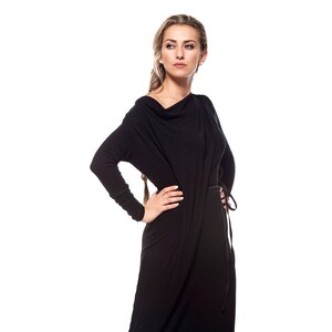 May include: A woman wearing a long black dress with a cowl neck and a belt. She is standing with her hand on her hip and looking to the side.